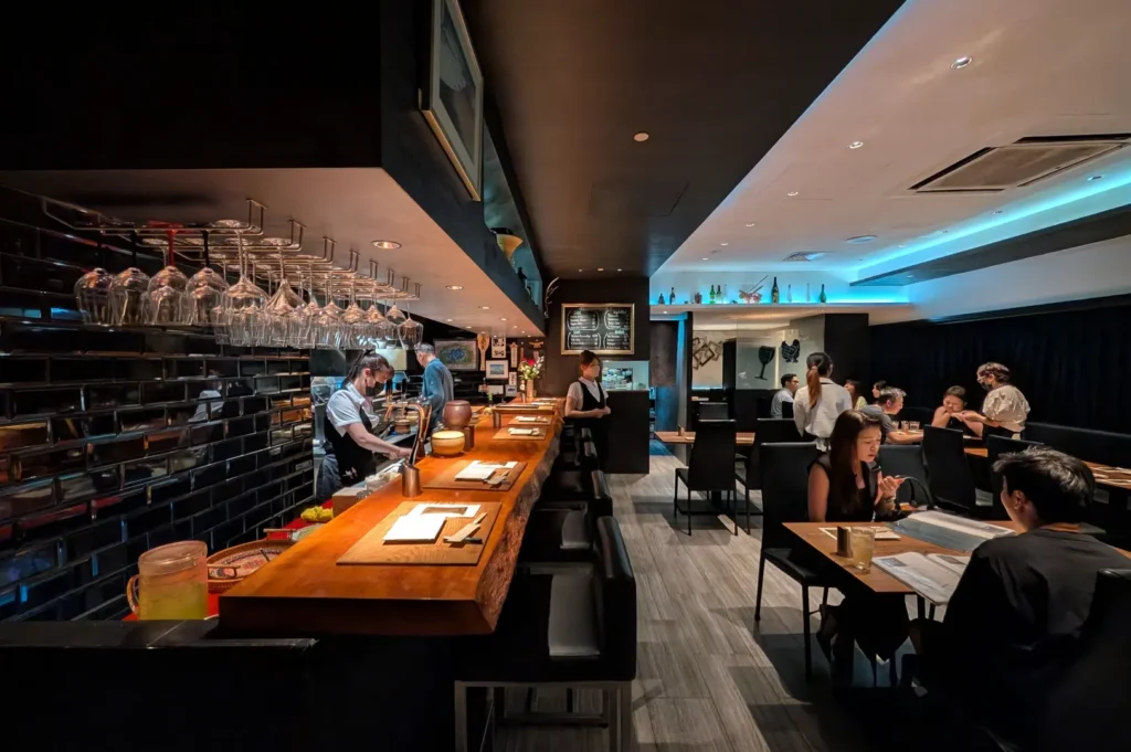 A wide, eye-level shot captures the sophisticated, modern interior of a dimly lit restaurant, representing the file "2-Best Yakitori in Cuppage Plaza Food Restaurants To Try.webp". On the left, a long, thick live-edge wooden bar counter is lined with black leather barstools, set against a dark brick-tiled wall beneath a massive row of hanging inverted wine glasses. Behind the bar, staff members in face masks and aprons work diligently, while a waitress in a white shirt stands ready near a blackboard menu in the center background. To the right, patrons sit at individual wooden tables on a grey wood-plank floor, dining under a sleek recessed ceiling feature illuminated by bright, cool blue neon accent lights.