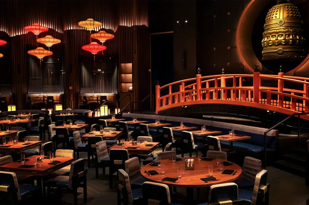 An eye-level shot captures the dramatic, dim, and upscale dining room of a restaurant, representing the file "2-Plaza Singapura vs Marina Bay Sands.webp". The expansive interior features a mix of neatly arranged square and round wooden tables set with black napkins, glassware, and small candles, paired with dark, low-backed chairs. The space is heavily defined by its striking Asian-inspired decor, notably a large, arched red wooden footbridge on the right side that spans across a section of the dining floor. Behind the bridge, a massive circular cutout in the wall showcases a monumental, hanging golden bell adorned with sculpted multi-faced deities, while the left side of the room is illuminated by glowing red and yellow traditional lanterns hanging against a backdrop of vertical wooden slats.