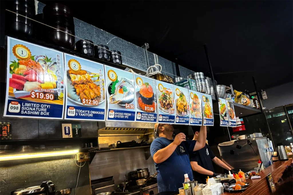 A low-angle, medium shot captures two kitchen staff members working diligently in an open kitchen under a long row of illuminated menu boards, representing the file "2-Sakedokoro Eizaburo Singapore.webp". The prominent, brightly colored menu signs hang overhead, showcasing various Japanese dishes with photos and prices, including a "Signature Sashimi Box," "Today's Omakase 5 Yakitori," "Hyogo Fresh Oyster," and an "Overflowing Ikura, Salmon & Negitoro Don." Below the signs, a chef wearing a dark blue t-shirt reaches up toward the menu structure, while a colleague next to him stands over a stainless steel food prep area stocked with squeeze bottles and ingredients. The busy kitchen environment is framed by stacks of black bowls on a shelf above the menus, warm under-counter lighting, and a dark wooden dining counter visible in the bottom-right corner.