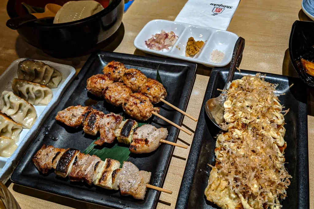 A high-angle, close-up shot captures an appetizing spread of traditional Japanese dishes on a light wooden table, representing the file "2-Shinjuku Restaurant Cuppage.webp". Centered on a long rectangular black plate are several skewers of yakitori, including glossy, grilled chicken meatballs (tsukune) and savory pork belly pieces interleaved with charred leeks (negima). To the right, a large platter showcases an okonomiyaki pancake generously drizzled with mayonnaise and savory sauce, covered in a mound of delicate, fluttering bonito flakes. On the left sits a plate of lightly browned gyoza dumplings, while small condiment dishes containing minced ginger, mustard, and salt sit neatly in the background under soft dining lights.