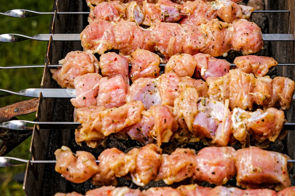 A close-up, high-angle shot captures raw, seasoned chunks of meat threaded tightly onto long metal skewers and arranged over a charcoal grill. The meat cubes are lightly coated with herbs and marinade, positioned side-by-side as they begin to cook over the hot coals below.
