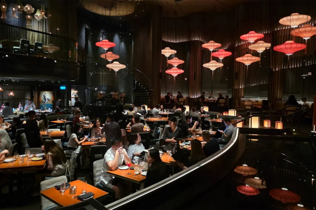 A wide, eye-level shot from an elevated vantage point captures the vast, dim, and energetic main dining room of an upscale Japanese restaurant, representing the file "2-The KOMA Experience.webp". Numerous wooden dining tables are tightly arranged across the main floor and filled with patrons socializing, looking at menus, and eating. The spectacular, high-ceilinged space features towering walls lined with dark, textured vertical slatted paneling and a curved staircase in the background. A grand, theatrical atmosphere is created by dozens of large, glowing traditional paper lanterns in alternating hues of vibrant red and soft cream, hanging at varied heights across the ceiling to cast a warm, dramatic light over the crowd.