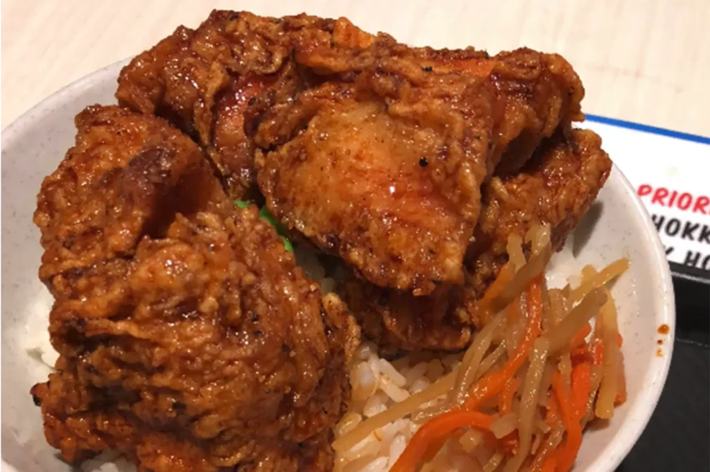 A close-up, high-angle shot captures a hearty Japanese rice bowl dish, representing the file "2-Yakitori Tsuyoshi Review What.webp". A speckled white ceramic bowl is piled high with several large pieces of golden-brown, crispy chicken karaage or fried chicken with a deeply textured, craggy crust. The chicken sits on a bed of steamed white rice, accented with a small portion of shredded orange carrots and pickled burdock root (kinpira gobo) in the lower right corner, creating a simple yet appetizing and satisfying meal presentation.
