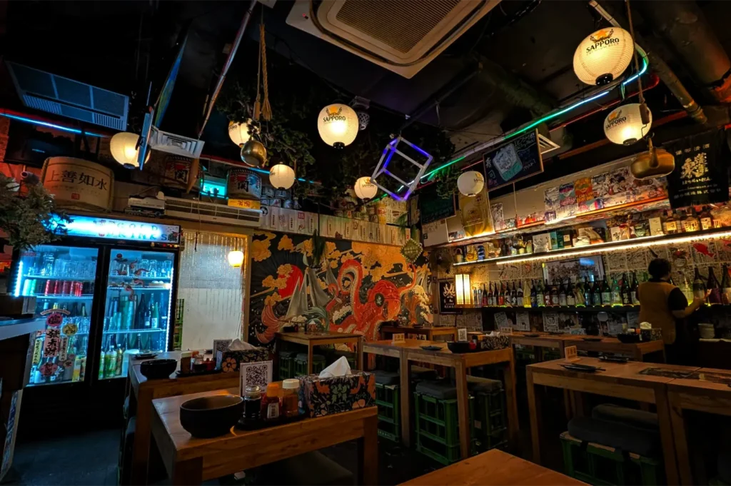 A wide, eye-level shot captures the vibrant, highly decorated interior of a Japanese izakaya, representing the file "2-Yorimichi Yakitori Izakaya Review.webp". The dining area features several thick wooden tables paired with stools made from green plastic crates topped with grey cushions, each set with condiments and tissue boxes. The back wall is covered with a dramatic, colorful mural depicting an octopus and ocean waves next to a bright blue-lit beverage refrigerator displaying cold sake and drinks. The lively, eclectic atmosphere is enhanced by a dark ceiling filled with hanging white Sapporo paper lanterns, neon tube accents, sake barrels, and extensive shelves packed with liquor bottles on the right wall where a staff member stands.
