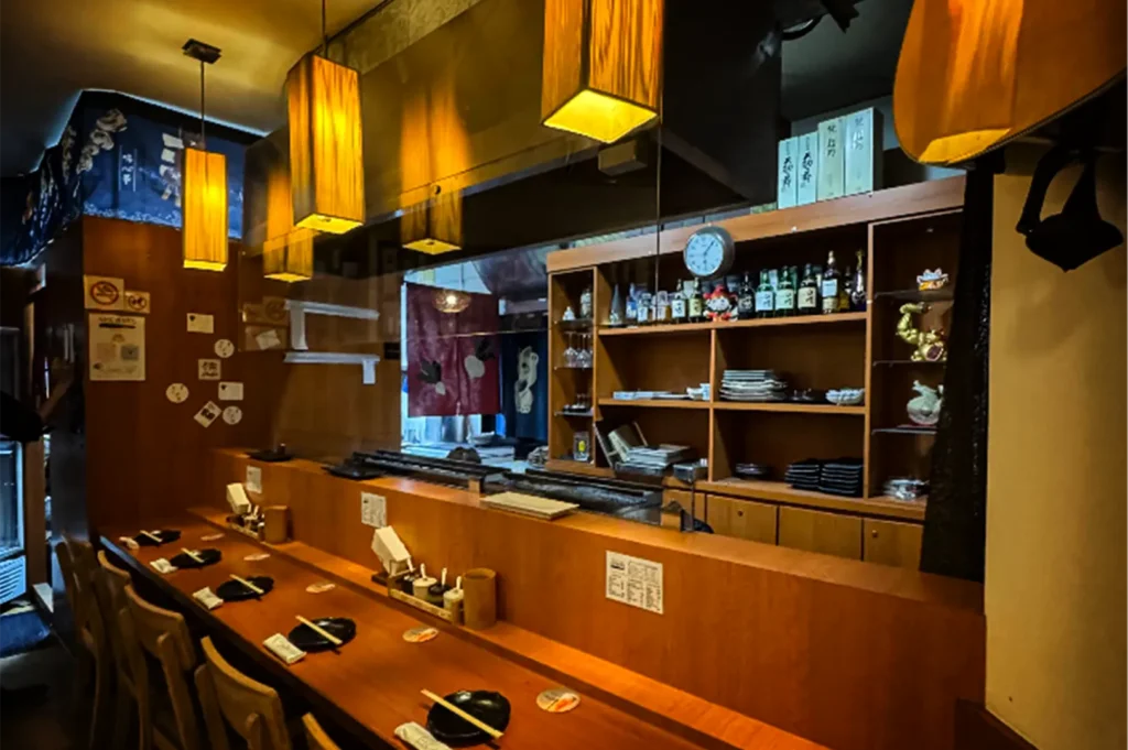 A wide, eye-level shot captures the quiet, empty interior of an intimate Japanese restaurant's dining counter, representing the file "3-Best Yakitori Near Plaza Singapura.webp". On the left, a long, smooth wooden counter is neatly set with dark ceramic plates, napkins, and chopsticks for individual diners, lined with high-backed wooden chairs. Above the counter, a series of rectangular, golden-glowing lantern-style pendant lights cast a warm ambiance over the space, which is separated from the back kitchen by a clear glass partition. The wooden shelves in the background are organized with sake and liquor bottles, stacks of plates, a wall clock, and decorative items like a waving lucky cat figure, while a traditional fabric noren curtain hangs in the rear doorway.