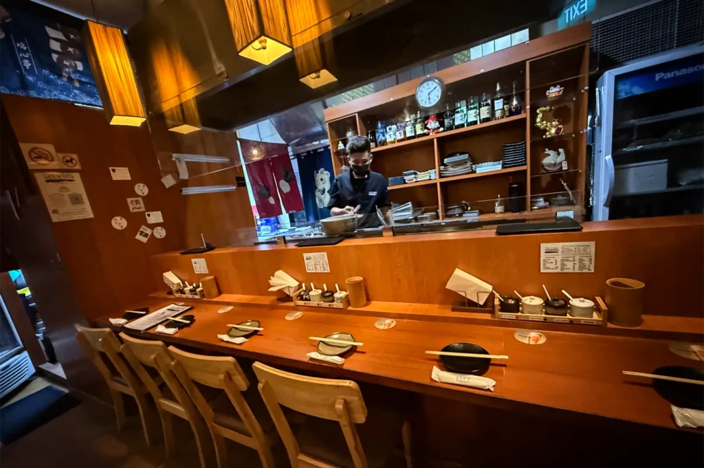 A wide, eye-level shot captures the warm, authentic counter seating area of a Japanese restaurant, representing the file "3-Best Yakitori in Cuppage Plaza Food Restaurants To Try.webp". A long, polished wooden bar counter spans the foreground, meticulously set with individual ceramic small plates, pairs of chopsticks resting horizontally, and sets of small condiment jars paired with napkins. Wooden dining chairs with open backs are lined up along the counter, facing a clear glass or acrylic partition that separates the seating from the kitchen service area. Behind the counter, a male chef wearing a black uniform and face mask works diligently at a grill or prep station beneath glowing, rectangular amber lanterns, with a large wooden shelf behind him displaying various liquor bottles, sake glasses, ceramic dishware, and decorative items.