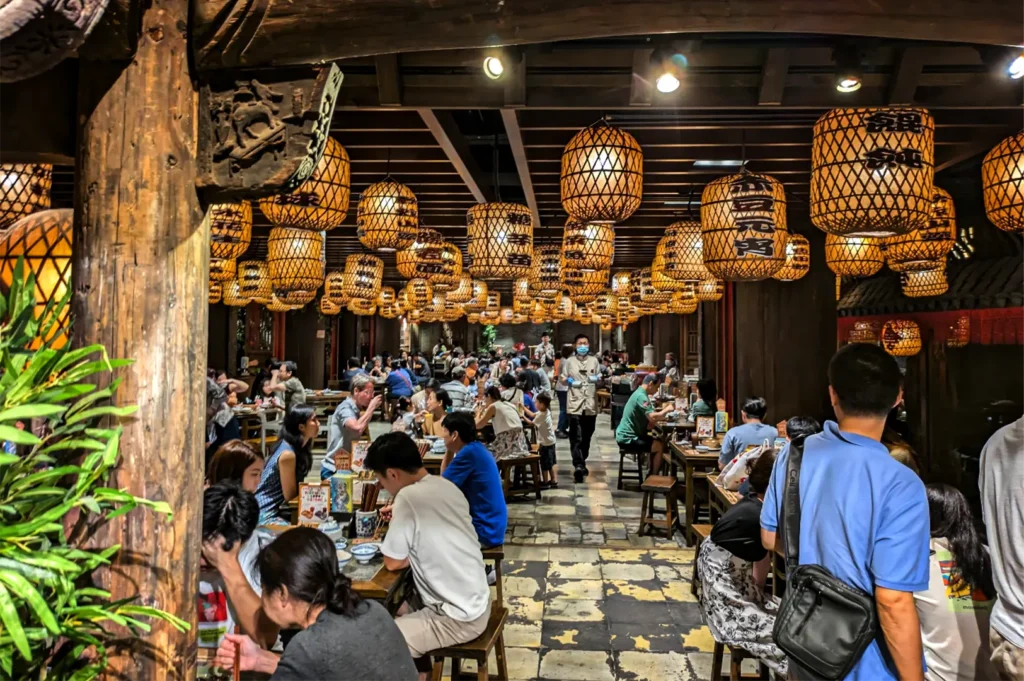 An eye-level shot captures a bustling, atmospheric restaurant filled with diners seated at wooden tables under a low ceiling densely packed with glowing orange lanterns. The interior features a rustic, traditional design with large wooden support beams—one of which is intricately carved in the foreground—and a stone-tiled floor. Dozens of large, woven-textured lanterns hang at varying heights, illuminating the crowded space with a warm, amber light and creating a sense of depth as they lead the eye toward the back of the room. A server wearing a face mask walks down the central aisle, navigating between the diverse groups of people eating and socializing, while a lush green plant on the far left adds a touch of natural color to the warm-toned environment.