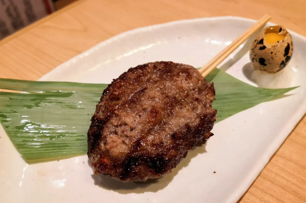 A close-up, high-angle shot captures a single grilled Japanese meatball skewer (tsukune) served on a long white ceramic plate, representing the file "3-Shinjuku Restaurant Cuppage_2.webp". The large, oval-shaped minced meat patty features a deeply browned, slightly charred surface with a juicy texture, resting atop a vibrant green bamboo leaf. Two wooden skewers extend from the top right of the patty, pointing toward a small, speckled quail egg with its top cracked open to reveal a bright yellow yolk, which sits nestled on a small mound of grated daikon radish. The dish is presented on a light wood tabletop under warm, soft restaurant lighting that highlights the savory details of the food.