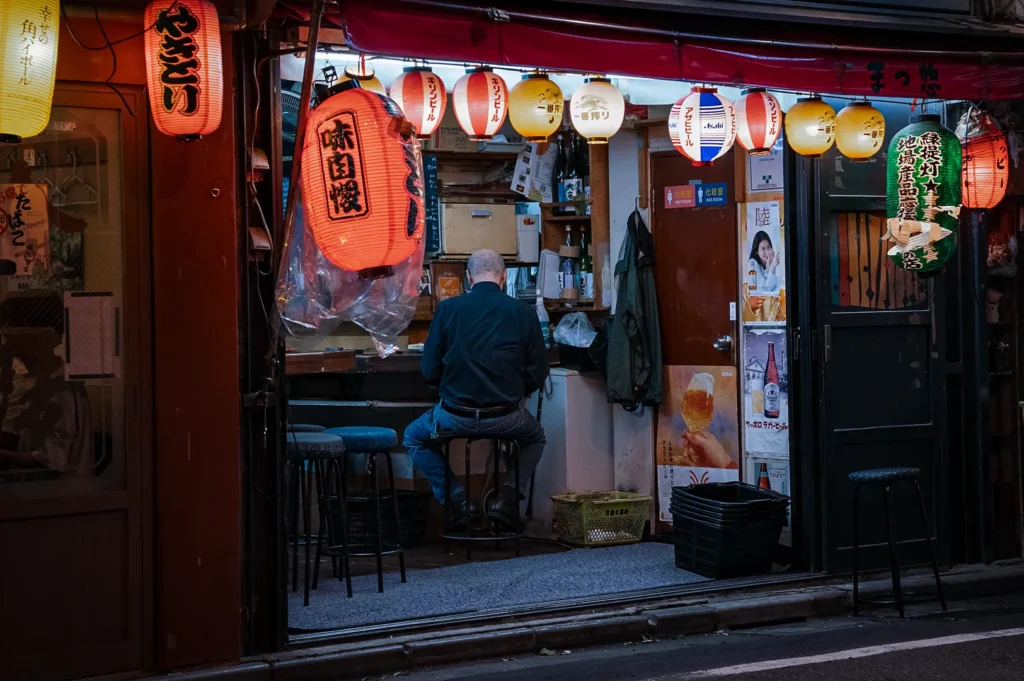 An eye-level shot captures a man in a black shirt seen from behind, sitting alone on a stool inside a narrow Japanese eatery at night. The cozy stall opens directly onto the street and is illuminated by a dense row of colorful, glowing paper lanterns hanging across the entrance.