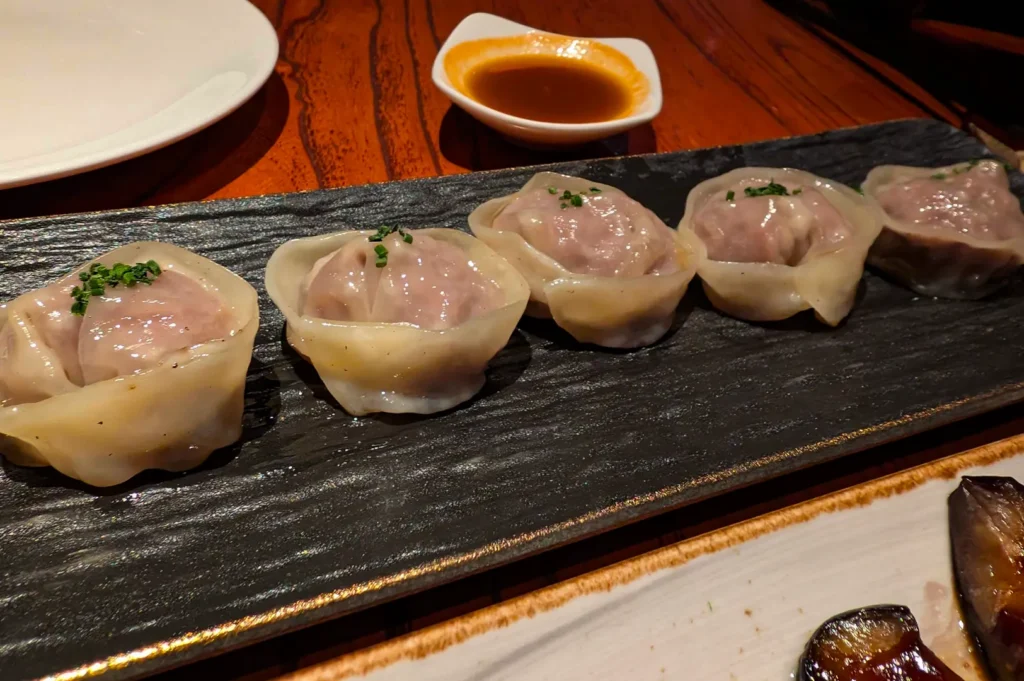 A close-up, high-angle shot captures a row of five beautifully crafted dumplings served on a rich wooden dining table, representing the file "3-The KOMA Experience.webp". Arranged in a neat horizontal line on a long, textured black slate platter, the plump dumplings feature translucent wrappers showing a savory meat filling, each delicately topped with a pinch of finely chopped green chives. In the softly blurred background, a small white ceramic dish holds a dark dipping sauce garnished with a touch of chili oil, while a portion of another plate containing cooked eggplant is partially visible in the bottom-right foreground, creating an appetizing fine-dining presentation.