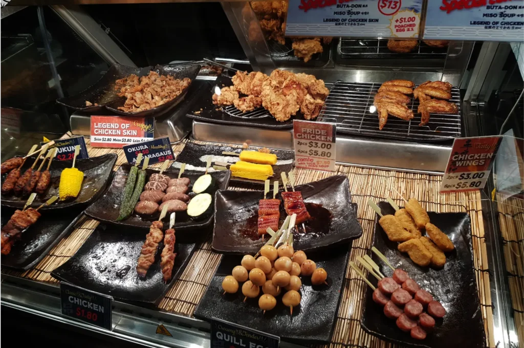 A slightly high-angle, medium close-up shot captures an organized food display case filled with various Japanese skewered and fried items, representing the file "3-Yakitori Tsuyoshi Review What.webp". Arranged on dark ceramic plates over a bamboo mat, the lower shelves feature an assortment of yakitori and sides on wooden skewers, including chicken, glazed unagi, quail eggs, sliced sausage, nuggets, shiitake mushrooms, okra, and corn on the cob. The raised metal shelves in the background showcase fried options, including a heap of crispy chicken skins labeled "Legend of Chicken Skin," a pile of golden-brown "Fride Chicken," and grilled chicken wings resting on wire racks, with clear price signs detailing the menu options throughout the display.