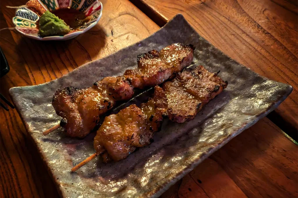 A high-angle, close-up shot captures two glistening, freshly grilled yakitori skewers served on a rustic rectangular ceramic plate, representing the file "3-Yorimichi Yakitori Izakaya Review.webp". The thick chunks of meat are skewered on wooden sticks, showing a savory, caramelized glaze with subtle charred edges from the grill that reflect the warm, overhead restaurant lighting. The square plate features an earthy, textured grey and brown glaze and rests on a polished wooden table with a visible grain. In the upper-left corner, a small, multi-colored decorative ceramic dish holds a dollop of bright green wasabi and a dark dipping sauce, completing the appetizing presentation.