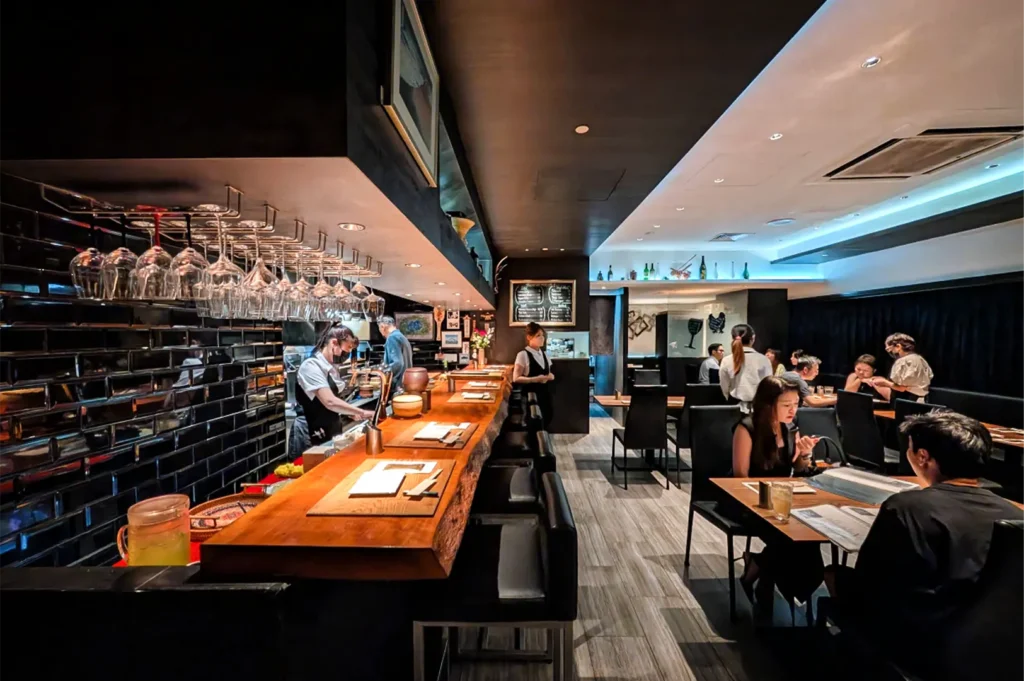 A wide, eye-level shot captures the sleek, modern interior of a dimly lit contemporary restaurant, representing the file "4-Best Yakitori Near Plaza Singapura.webp". On the left, a long, thick wooden bar counter with a live-edge design features a row of empty black upholstered barstools, set against a glossy black-tiled wall where dozens of wine glasses hang upside down from overhead racks. Behind the counter, kitchen staff in uniform are seen preparing orders. To the right, a spacious dining area with light grey wood-plank flooring is filled with several groups of patrons seated at dark wooden tables with black leather chairs, all illuminated by cool white recessed ceiling lights and soft under-counter ambiance.