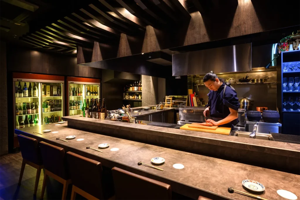 A wide, eye-level shot captures the sleek, upscale interior of a modern Japanese restaurant's open kitchen and counter area, representing the file "4-Best Yakitori in Cuppage Plaza Food Restaurants To Try.webp". In the foreground, an L-shaped, smooth concrete or stone dining counter is lined with elegant dark leather chairs, neatly set with pairs of chopsticks and small ceramic plates. Behind the counter, a male chef with his hair tied back in a bun stands at a wooden cutting board under a stainless-steel exhaust hood, meticulously preparing food. The sophisticated, dimly lit ambiance is highlighted by an intricate wooden slatted ceiling design and a pair of large, brightly lit glass-door refrigerators in the background that showcase an extensive collection of sake and wine bottles.