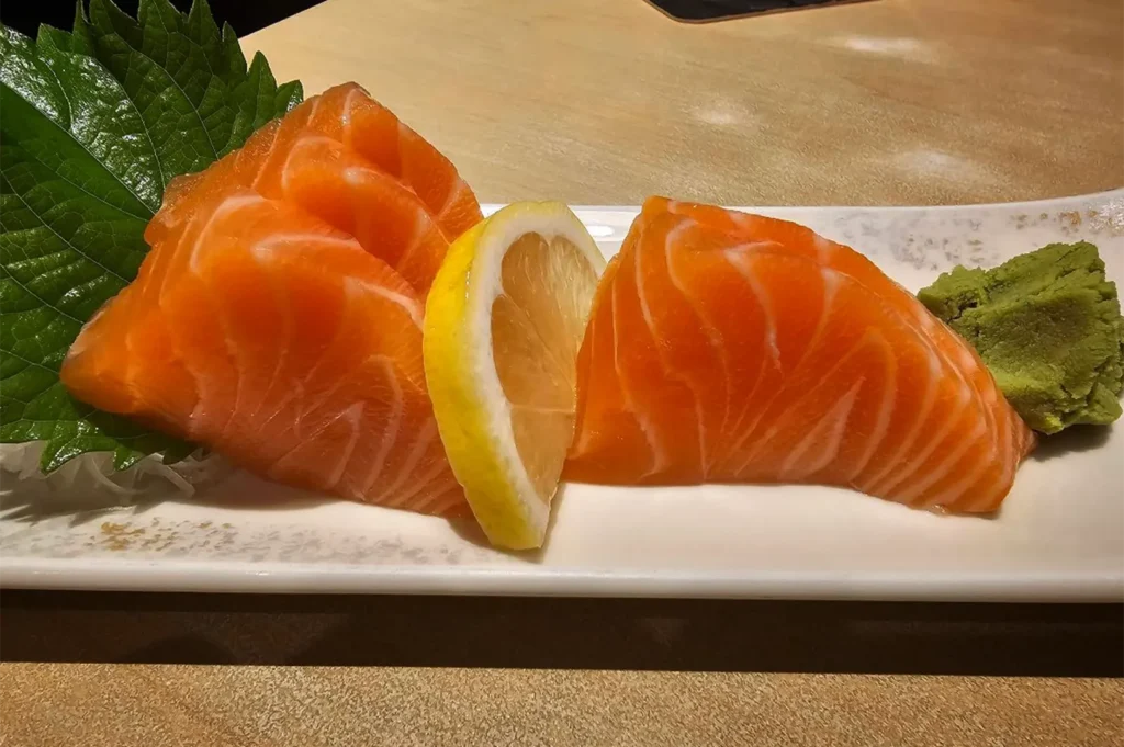 A high-angle, close-up shot captures a fresh and vibrant serving of salmon sashimi on a long, narrow white ceramic platter, representing the file "3-Shinjuku Restaurant Cuppage.webp". The dish features thick, triangular slices of glossy, bright orange salmon showcasing distinct white marbling, neatly arranged into two distinct piles. A half-wheel slice of juicy yellow lemon separates the two portions of fish, while a large, jagged green shiso leaf sits on the left over a bed of shredded daikon radish, and a textured dollop of pale green wasabi paste rests on the far right. The plate sits angled across a light-colored wooden table, illuminated by warm overhead dining lights that accentuate the fresh, appetizing appearance of the seafood.