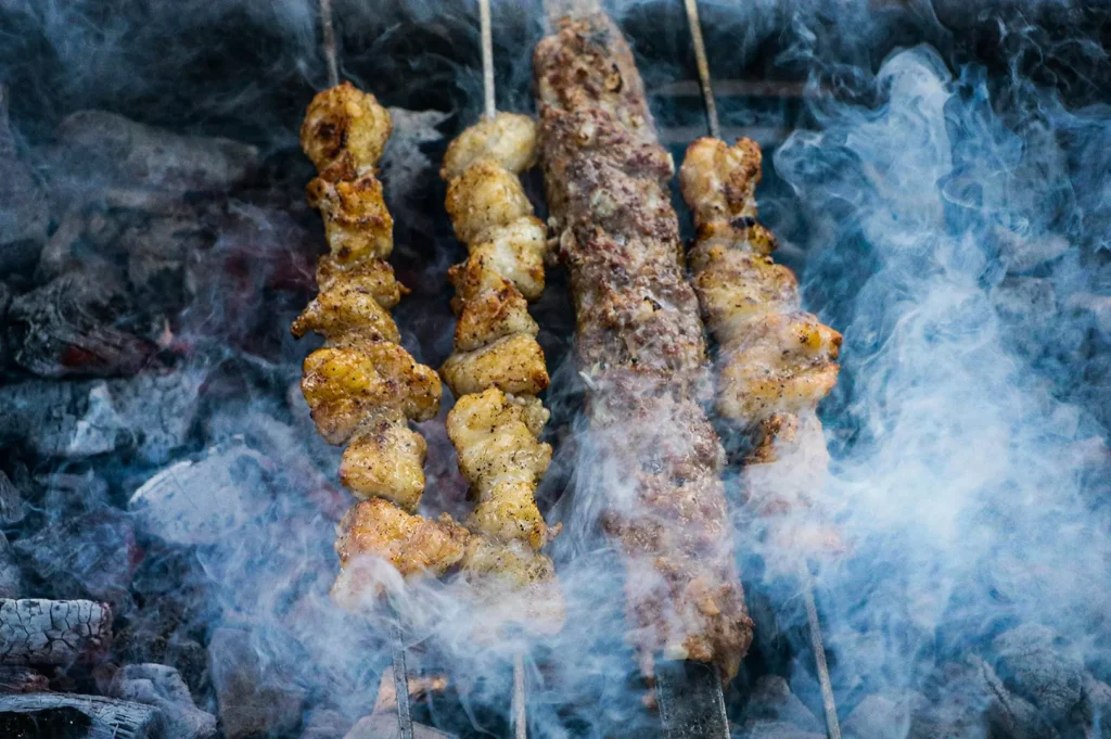 A close-up, top-down shot captures four metal skewers of meat chunks and ground minced meat grilling over hot charcoal. Thick plumes of white smoke billow up from the embers below, partially obscuring the kebabs as they sizzle.