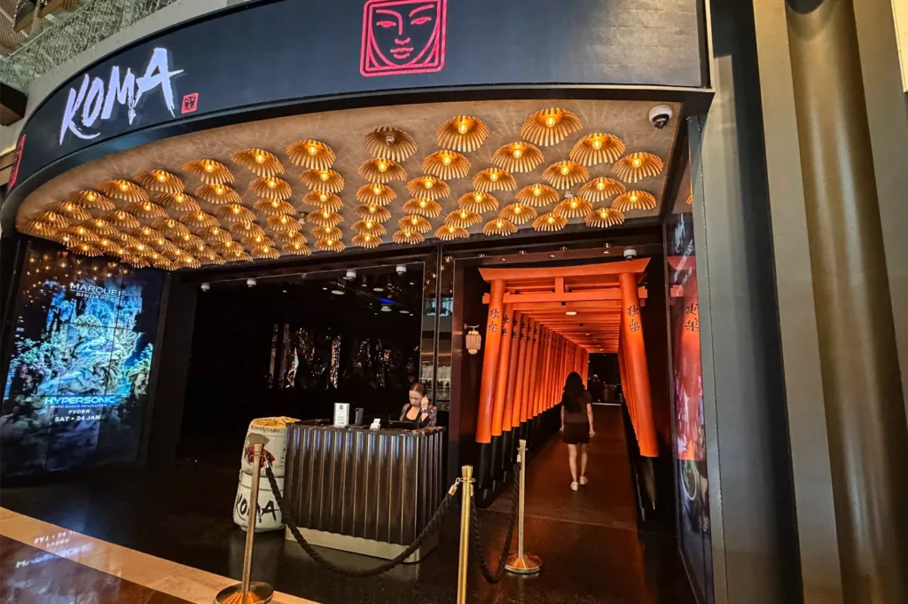 A wide, eye-level shot captures the dramatic, grand entrance of a luxury restaurant located inside a modern commercial building, representing the file "4-The KOMA Experience.webp". The exterior features a prominent curved black sign overhead displaying the stylized restaurant name "KOMA" in white brush-stroke lettering next to a glowing pink icon of a stylized face. Beneath the entrance, a magnificent ceiling is densely lined with dozens of small, inverted straw hats glowing with warm, golden light. A black fluted reception desk stands at the entrance behind a stanchion rope barrier, where a staff member works, while to the right, a customer walks down a long, striking tunnel walkway constructed from closely aligned, bright orange traditional Japanese torii gates that lead into the dim restaurant interior.