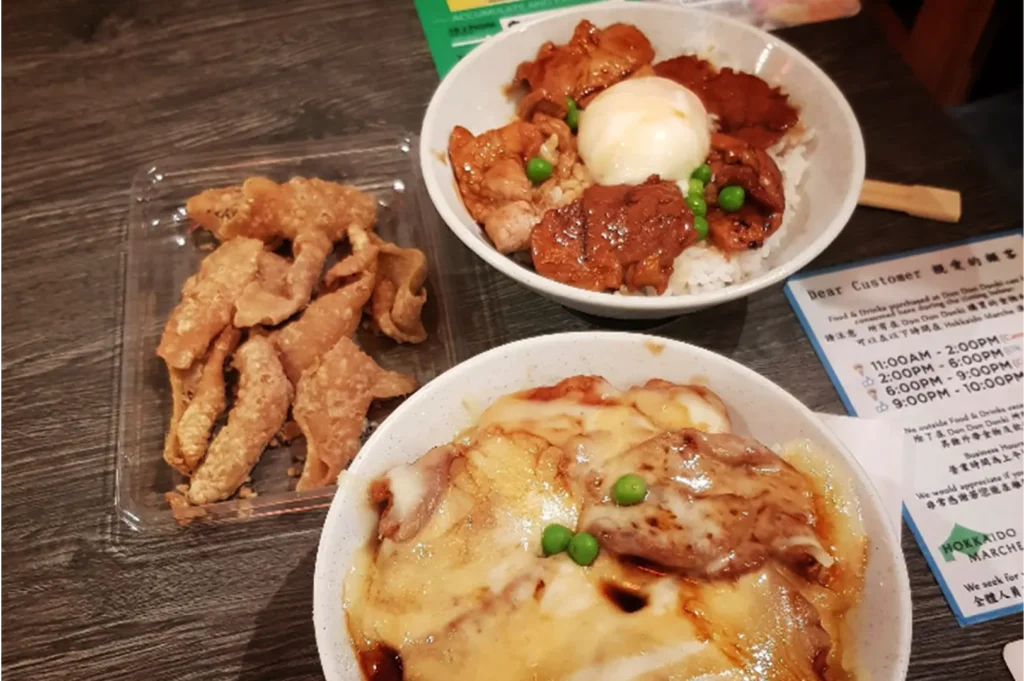 A high-angle, medium close-up shot captures a delicious Japanese meal layout on a dark wooden table, representing the file "4-Yakitori Tsuyoshi Review What.webp". In the foreground, a large ceramic bowl features a pork rice bowl covered in a generous layer of melted cheese, drizzled with sweet soy glaze and garnished with green peas. Positioned just behind it on the right is a traditional "buta-don" pork bowl topped with a soft-boiled egg, glazed pork slices, and green peas, while a clear plastic container filled with crispy fried chicken skins sits on the left. In the background, a small informational notice card for "Hokkaido Marche" is partially visible, rounding out the casual dining setting.