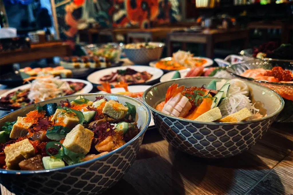 A close-up, eye-level shot captures a lavish spread of vibrant Japanese dishes arranged across a dark wooden table, representing the file "4-Yorimichi Yakitori Izakaya Review.webp". In the foreground, two large ceramic bowls with a textured, scale-like blue and white pattern are filled with colorful ingredients; the left bowl contains a chirashi-style mix of cubed tamago (egg omelet), cucumbers, ikura (salmon roe), and seaweed, while the right bowl neatly showcases slices of fresh salmon, shrimp, squid, and shredded daikon radish. The background falls into a soft blur, revealing a crowded feast of additional platters including sashimi, skewers, and side dishes, set within a dimly lit restaurant where a colorful wall mural is faintly visible in the distance.