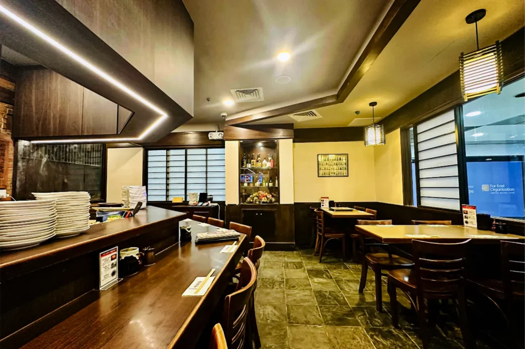 A wide, eye-level shot captures the quiet, empty dining room of a traditional Japanese restaurant, representing the file "5-Best Yakitori Near Plaza Singapura.webp". On the left, a long, dark-stained wooden counter angles through the space, set with menu cards, small cups, and wooden chopsticks, while a large stack of white plates rests on a raised ledge under an overhanging geometric ceiling structure illuminated with a strip of warm LED lighting. The main dining floor features dark, textured stone-look tiles and is arranged with several wooden tables and matching slatted-back chairs. In the background, beige walls are complemented by dark wood paneling, featuring a recessed display cabinet filled with liquor bottles, windows with traditional white shoji-style screens, and a large glass window on the right looking out into a brightly lit office corridor displaying a "Far East Organization" logo.