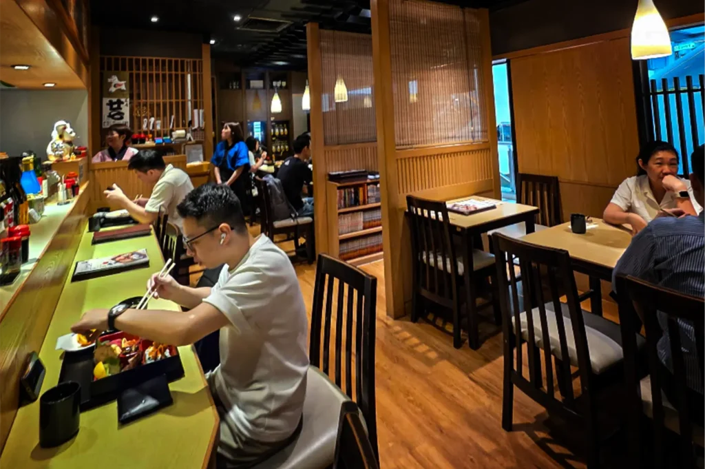 A wide, eye-level shot captures the bustling, warmly lit interior of a Japanese restaurant filled with patrons dining, representing the file "5-Best Yakitori in Cuppage Plaza Food Restaurants To Try.webp". In the foreground on the left, a man wearing glasses and a white t-shirt sits at a long, light-green counter, using chopsticks to eat from a bento box, while another diner is seated further down the counter. To the right, separate wooden dining tables and chairs are arranged on a polished wood floor, with a couple chatting at one of the tables. The traditional izakaya-style ambiance is enhanced by wooden wall paneling, a large wooden privacy screen separating seating areas, hanging teardrop pendant lights, and restaurant staff moving between tables in the background.