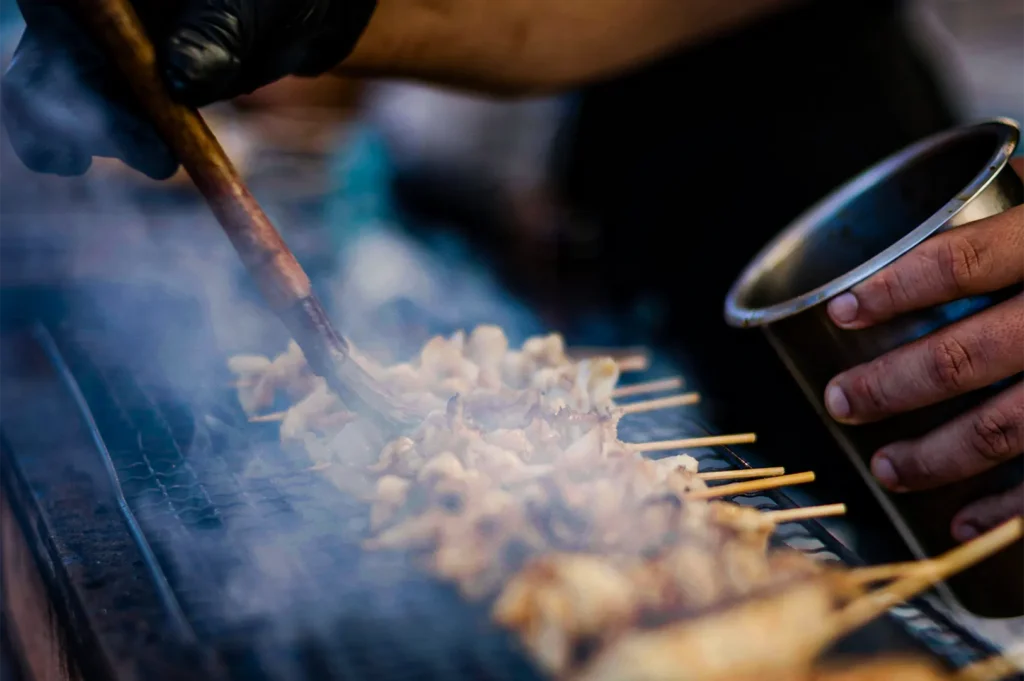 A close-up shot captures a vendor using a long wooden brush to baste rows of small meat skewers cooking on a charcoal grill. Thick wisps of smoke rise from the grate while the vendor holds a stainless steel cup of sauce in their other hand.