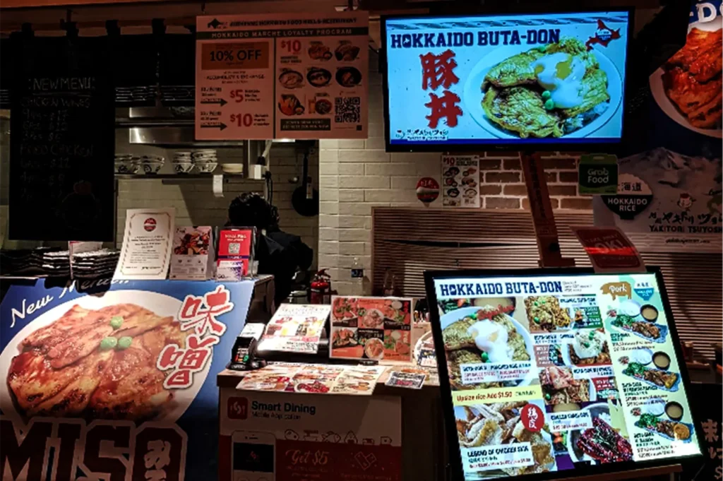 A medium, eye-level shot captures the busy, brightly lit ordering counter of a casual Japanese food stall, representing the file "6-Best Yakitori Near Plaza Singapura.webp". The storefront is heavily decorated with promotional signage, featuring a large, colorful banner on the bottom left advertising a "New Menu... Miso" pork dish, a smaller "Smart Dining" mobile app standee in the center, and a large illuminated menu board on the right detailing various "Hokkaido Buta-Don" pork and chicken options. Overhead, a digital television screen dynamically displays a close-up photo of a pork bowl topped with a soft-boiled egg under the words "Hokkaido Buta-Don," positioned next to hanging printed signs outlining a "Hokkaido Marche Loyalty Program." In the background, a staff member in a dark uniform works in the kitchen behind a white-brick tiled wall holding stacks of bowls.