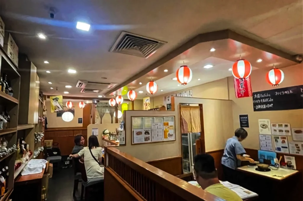 A wide, eye-level shot captures the brightly lit, casual dining room of an authentic Japanese restaurant, representing the file "6-Best Yakitori in Cuppage Plaza Food Restaurants To Try.webp". The cozy interior is partitioned by low wooden walls, creating intimate dining spaces where patrons are seated at various tables, including a couple chatting on the left and a diner reading a menu in the foreground. On the right, a staff member in a blue shirt prepares a table beneath a small blackboard menu and informational posters affixed to the wall. The traditional, lively izakaya ambiance is highlighted by a long row of glowing red-and-white paper lanterns hanging from the ceiling, leading down a narrow walkway flanked by open wooden shelving filled with bottles and decorative items on the far left.